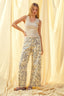 Woman wearing a white sleeveless top and patterned pants standing against a soft, light-colored curtain.