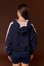 A woman viewed from behind wearing a navy blue hoodie with light pink contrast panels on the hood lining and sleeves, paired with navy shorts, against a solid dark brown studio background.
