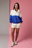 Woman wearing a blue and white color-blocked sweater with white shorts on a pink background