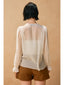 Person wearing a sheer beige blouse over a white shirt with brown shorts against a beige background