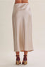 A model's lower body wearing a high-waisted, midi-length satin slip skirt in a light cream color, paired with dark burgundy high-heeled sandals, against a solid light beige background.