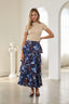 A woman modeling a beige short-sleeve mock-neck top and a navy blue tiered maxi skirt with a colorful floral and paisley print, standing in a bright, white-walled room with arches.