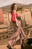 A woman in a rustic western-style outfit walking outdoors in a dusty, desert-like setting with mountains and wooden fences in the background. She is wearing a red top, a long red-and-white gingham skirt, white stockings, and dark boots, while holding a woven basket.