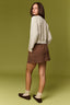 A woman stands in profile wearing a cream cable-knit cardigan, a brown mini skirt, cream socks, and dark brown flats against a solid olive green studio background.