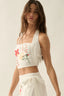 Woman wearing a white sleeveless top with floral embroidery and white pants on a plain background