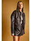A woman modeling a dark brown faux-leather jacket with a pointed collar, button front, and large front pockets, set against a solid warm brown background.