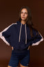 A woman modeling a navy blue hoodie with contrasting light pink sleeves and hood drawstrings, paired with matching navy athletic shorts, against a solid dark brown studio background.