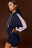 A woman modeling a navy blue color-blocked hoodie with light pink sleeves and matching navy athletic shorts against a solid dark brown studio background.
