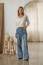 Woman wearing a white top and blue jeans standing in a room with a wooden stool and vase.