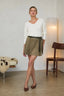 A woman stands in a minimalist room wearing a white long-sleeve V-neck top, an olive green wrap-style mini skirt with a black belt, and brown pointed-toe flats.