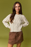 A woman wearing a cream cable-knit crewneck sweater and a brown drawstring mini skirt, standing against a solid olive green studio background.