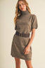 A woman modeling a taupe brown short-sleeve knit turtleneck dress with a wide dark brown belt featuring a gold buckle, set against a plain beige background.