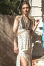 A woman wearing a bohemian-style two-piece set featuring a halter crop top and a matching high-slit maxi skirt, standing outdoors in front of a wooden fence and stone wall.
