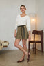 A woman models a white long-sleeve knit top and olive green suede shorts with a dark green tie belt, paired with brown leather slingback flats, in a minimalist room setting.