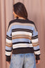 A woman shown from the back wearing a striped knit sweater with horizontal bands of dark brown, light blue, beige, and white, paired with light-wash blue jeans against a soft pink curtain backdrop.