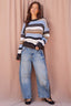 A woman modeling a striped knit sweater with blue, brown, black, and white horizontal stripes, paired with wide-leg light-wash jeans and black flats, standing against a dusty pink fabric backdrop.
