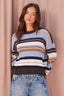 A woman wearing a striped knit sweater with horizontal bands of light blue, white, brown, and black, paired with light-wash blue jeans, standing against a dusty pink curtain backdrop.