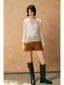 Woman wearing a white blouse, brown shorts, and black knee-high boots against a beige wall.