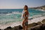 Woman in a bikini standing on rocks by the ocean holding a magazine.