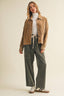 A woman modeling a brown corduroy jacket over a white turtleneck, paired with wide-leg dark green corduroy pants and white sneakers, against a plain beige studio background.