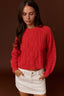 A woman wearing a bright red cable-knit sweater and a white mini skirt, standing against a dark brown studio background.