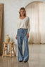 Woman wearing a white sweater and blue jeans standing in a room with a wooden stool and decorative vase.