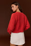 A woman wearing a bright red cropped knit sweater with textured cable detailing on the sleeves, paired with a white mini skirt, photographed against a solid dark brown studio background.
