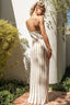 Woman in a white pleated dress standing outdoors with plants and a wall in the background