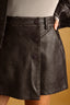 Close-up of a black faux-leather mini skirt with a button-front closure and side pockets, worn with a ribbed top against a warm tan background.