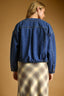 A woman shown from the back wearing a cropped blue denim jacket over a beige and brown plaid skirt, set against a solid warm brown studio background.