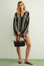 Woman wearing a black and white striped dress holding a black handbag against a plain background