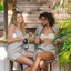 Two women sitting outdoors, one holding a bottle and the other a drink, with a wooden structure and plants in the background.