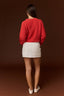 A woman shown from behind wearing a bright red textured knit sweater and a white mini skirt, standing against a solid dark brown studio background.