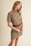 A woman modeling a taupe knit mock-neck dress with short sleeves and a matching dark brown belt with a silver buckle, set against a plain beige background.