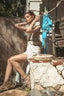 A woman wearing a patterned sleeveless dress and strappy high-heeled sandals sits on a stone ledge in an outdoor backyard setting. A blue bikini is hanging on a clothesline behind her, with a woven basket on a brick ledge in the foreground.