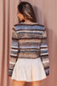 A woman shown from the back wearing a fuzzy striped knit sweater with horizontal bands of blue, brown, beige, and white, paired with a white skirt, set against a soft pink curtain backdrop.