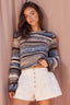 A woman wearing a fuzzy striped knit sweater in shades of blue, brown, beige, and gray, paired with white ruffled shorts, posing against a dusty pink fabric backdrop.