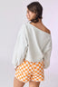 Woman wearing a light gray off-shoulder sweater and orange checkered shorts on a white background
