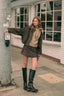 A woman stands on a city sidewalk wearing a dark brown leather jacket, a beige knit sweater, a matching brown mini skirt, and black knee-high boots, posing next to a gray utility pole in front of a white brick building.