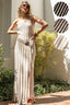Woman in a long, light-colored dress standing outdoors with plants and a white wall in the background.