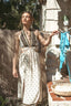 A woman stands outdoors in a sunny backyard wearing a sleeveless halter maxi dress with a dark brown sequined bodice and a cream patterned skirt. She is holding up a blue bikini top on a clothesline next to a rustic stone wall.