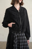 A person wearing a black cropped zip-front jacket with a ribbed collar, cuffs, and hem, paired with a black-and-gray plaid skirt, against a plain light beige wall.