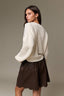 A woman wearing a cream waffle-knit cropped sweater and a dark brown flared skirt, photographed against a plain warm gray studio background.