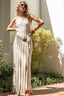 Woman in a long, light-colored dress standing outdoors with greenery and a white wall in the background.
