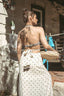 A woman stands outdoors in a sunlit courtyard wearing a white halter sarong with a brown geometric pattern, holding a woven straw bag. The back of the halter top has a brown and silver patterned design.