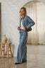 A woman models a matching light-wash blue denim set consisting of a cropped button-up jacket and wide-leg pants with brown leather side panels, styled with a white top and a brown crossbody bag in a rustic indoor setting.