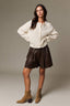 A model posing in a cream ribbed knit cardigan, a dark brown faux-leather mini skirt, and tan suede ankle boots against a neutral gray studio backdrop.