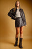 A woman models a dark brown faux-leather jacket and matching mini skirt set, styled with a light gray top and dark brown knee-high boots, against a warm brown studio backdrop.