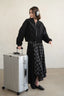 A woman wearing a black bomber jacket, a black and gray plaid skirt, and black loafers stands next to a silver aluminum hardshell suitcase, wearing gray over-ear headphones against a neutral beige background.