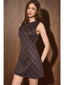 A woman modeling a sleeveless, knee-length A-line dress with a dark plaid pattern in black, burgundy, and brown, posed against a dark wood-paneled wall.
